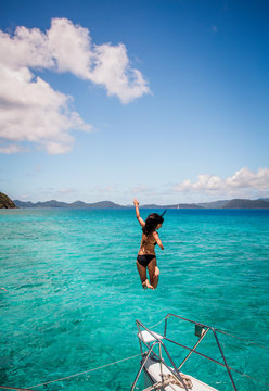 BRITISH VIRGIN ISLANDS, CARIBBEAN. Woman in black bikini jumps off the bow of a catamaran into teal Caribbean water.