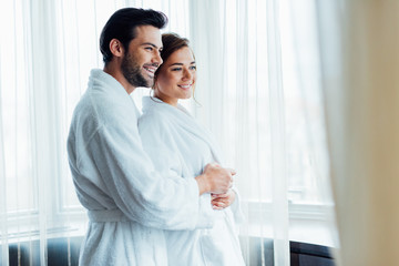 selective focus of cheerful man hugging happy woman in white bathrobe in hotel