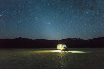 ALVORD DESERT, HARNEY COUNTY, OR, USA. A white van is parked in a giant dried lake bed at night under a starry, Milky Way sky with what appears to be a halo of light around the van.