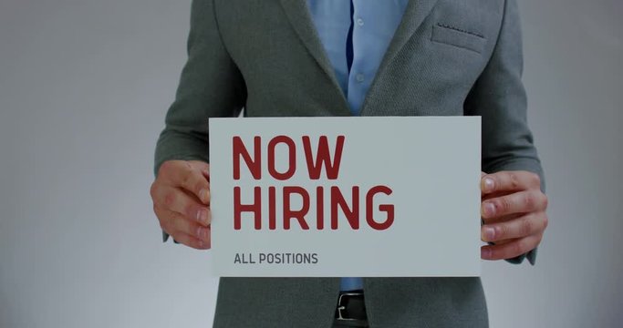 FIXED Handsome Caucasian HR manager wearing suit holding recruitment sign saying Now hiring. Light grey background. 4K UHD