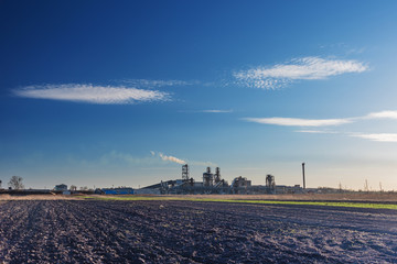 Industrial spring landscape with a view of a wood processing plant with high smoking pipes, against a background of blue sky and agricultural fields.