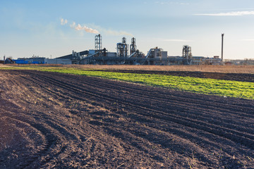 Fototapeta na wymiar Industrial spring landscape with a view of a wood processing plant with high smoking pipes, against a background of blue sky and agricultural fields.