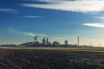 Fototapeta na wymiar Industrial spring landscape with a view of a wood processing plant with high smoking pipes, against a background of blue sky and agricultural fields.