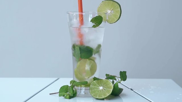 orange straw stirs fresh mojito in tall glittering glass near lime halves and mint leaves on white table slow motion