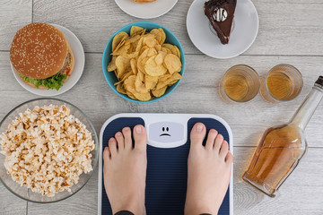 Woman standing on scales surrounded by different food and alcohol after party indoors, top view