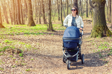 Fototapeta na wymiar Beautiful young adult woman walking with baby in stroller through forest or park on bright sunny day. Healthy lifestyle and children healthcare. Happy childhood and parenthood