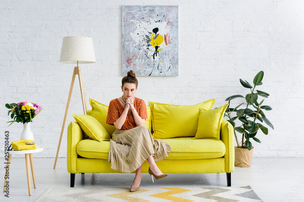 Wall mural pensive elegant young woman sitting with crossed legs and clenched hands on yellow sofa in living ro