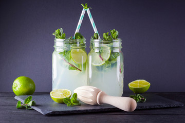 Mojito and mint on black background. Refreshing cocktail with lime and fresh mint on slate board. Two jars with handles with a summer drink with citrus and ice. Manual wooden juicer and mojito
