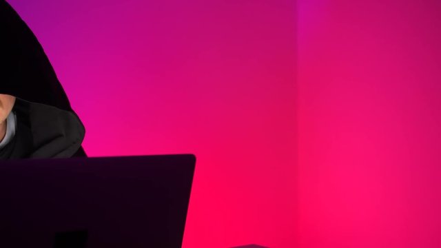 A young hacker in a black gown is working at a computer on a cyber attack in the office. Anonymous user commits cybercrime
