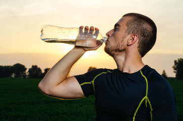 guy brunette in gym clothes. training in the field, drink clean water with bottle. in the Park at sunset.