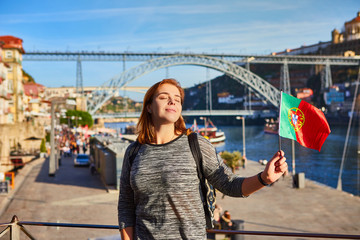 Young woman traveler standing back with portuguese flag, enjoying beautiful cityscape view on Douro river, bridge and boats during the morning light in Porto, Portugal