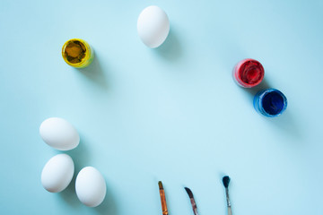 A set of white clean eggs, blue, yellow and red paints and brushes on bright background top view, flat lay. Easter symbols. Easter eggs, painting eggs, easter illustration image.  