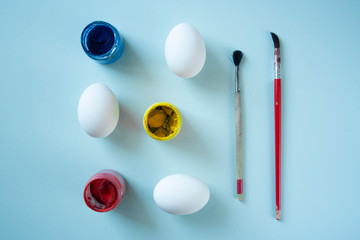 A set of white clean eggs, blue, yellow and red paints and brushes on bright background top view, flat lay. Easter symbols. Easter eggs, painting eggs, easter illustration image.  