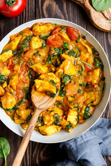 Top view on Bombay chicken curry with spinach, tomato and onion in a white frying pan on a kitchen wooden table. Indian traditional cuisine.