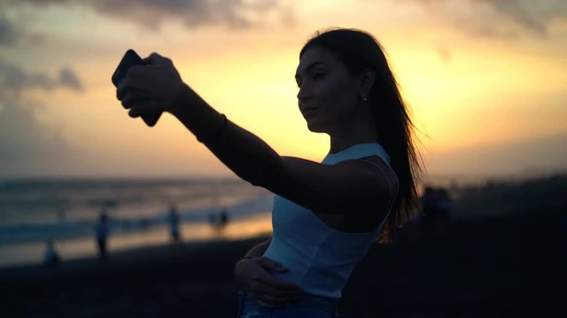 Young woman taking selfie photo with cellphone on beach during sunset