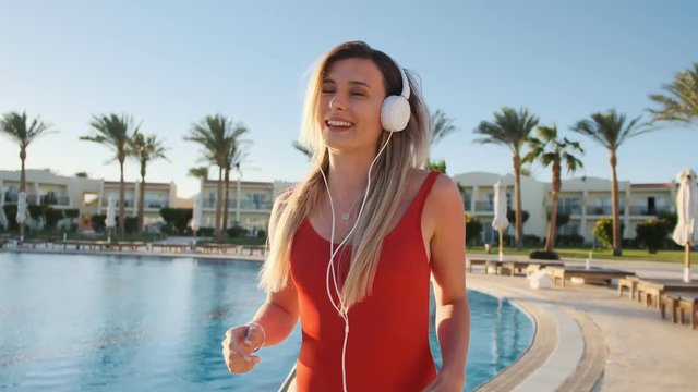 Beautiful woman in red swimsuit and headphones dancing near clear blue swimming pool background, have fun and listening music on smartphone using app. Bikini girl having good time on summer holiday.