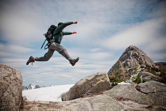 A backpacker jumps between two granite rocks over a patch of snow en route to Needle Peak, BC, Canada.