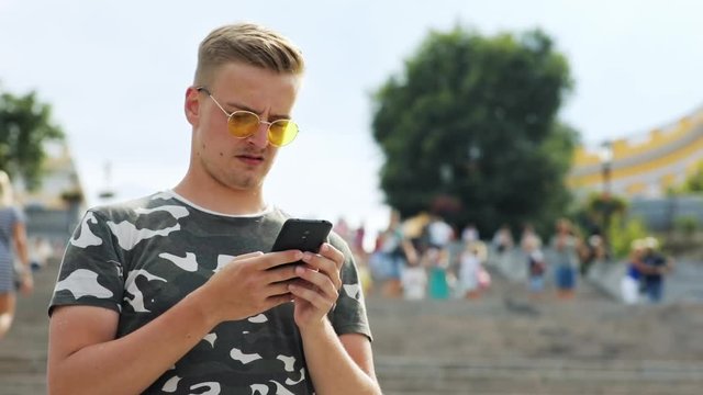 Handsome blonde man wearing sunglasses texting on smartphone on the large stairway. Cityscape at sunlight.