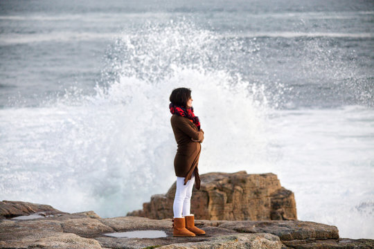 Woman standing on rocky coastline with waves crashing behind