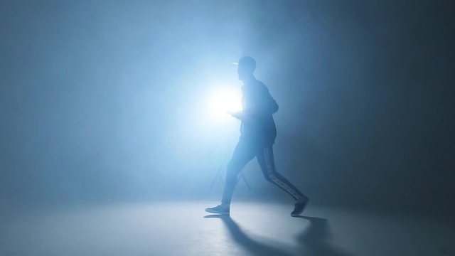 Sporty modern style hip-hop dancer dressed in urban style wear shows his dance on blue studio background, slow motion.