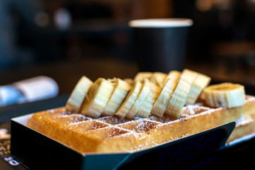 Fresh square Belgian waffle with cut banana topping and sugar powder for breakfast on dark background.