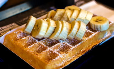 Fresh square Belgian waffle with cut banana topping and sugar powder for breakfast on dark background.