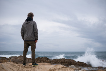 Traveler on the rocks near the sea looking far away at horizon. Rocky Atlantic Ocean Coastline and Stormy Weather. Handsome young caucasian tourist man in casual clothes outdoors on the nature
