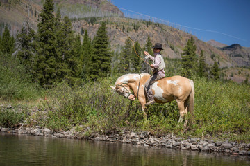 A man fishes from the back of a pinto horse in cowboy hat in Montana backcountry near Yellowstone.