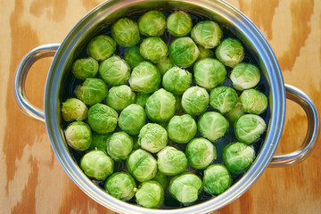 Top view on stainless steel cooking pot with water and bunch of fresh harvested green brussels sprouts before cooking on the wooden table. Light and healthy vegetarian or vegan dish, full of vitamins.
