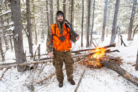A male hunter in camouflage with a blaze orange vest on holds his rifle next to a fire he started when the weather turned colder during an elk and deer hunting trip in Northwest Montana.