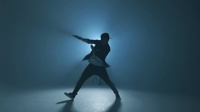 Portrait of a young man dancer in casual street wear dancing funky hip hop on isolated studio neon blue background, slow motion