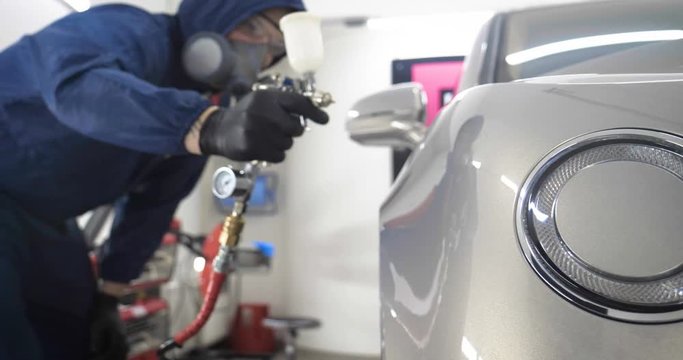 In slow motion, a Man (Man) professional in working uniform with spray guns in his hands and a respirator in protective glasses puts paint (ceramics) on the car on different parts of the car.