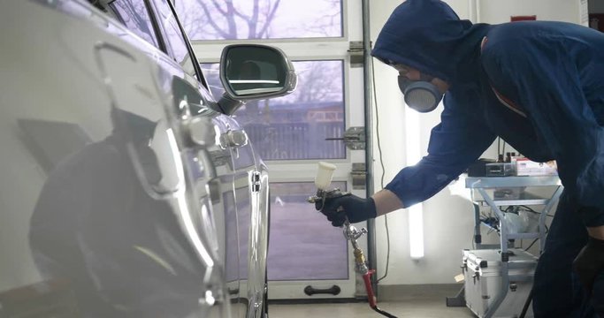 In slow motion, a Man (Man) professional in working uniform with spray guns in his hands and a respirator in protective glasses puts paint (ceramics) on the car on different parts of the car.