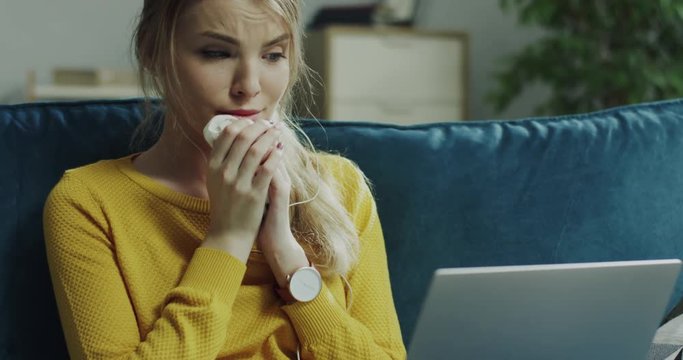 Portrait shot of the pretty blonde young woman watching sad romantic movie on the laptop computer and crying in the napkin on the sofa in the living room.