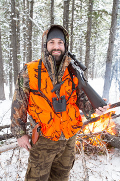 A male hunter in camouflage with a blaze orange vest on holds his rifle next to a fire he started when the weather turned colder during an elk and deer hunting trip in Northwest Montana.