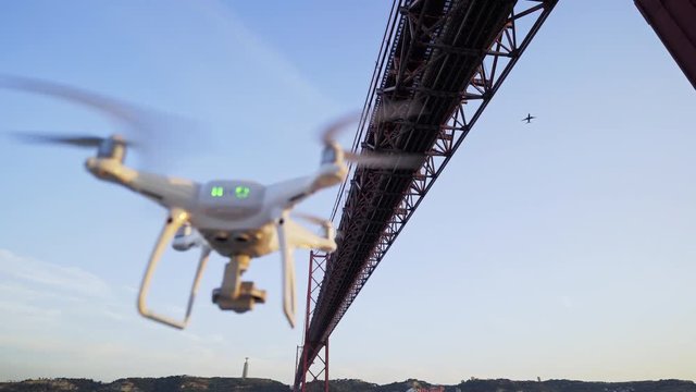 Closeup of modern white drone with green lights flying in no fly zone under suspension bridge near airport. Lisbon, Portugal