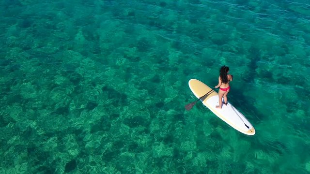 Aerial drone bird's eye view video of attractive fit young woman practicing stand up paddle board or SUP in tropical caribbean sapphire crystal clear sandy exotic beach