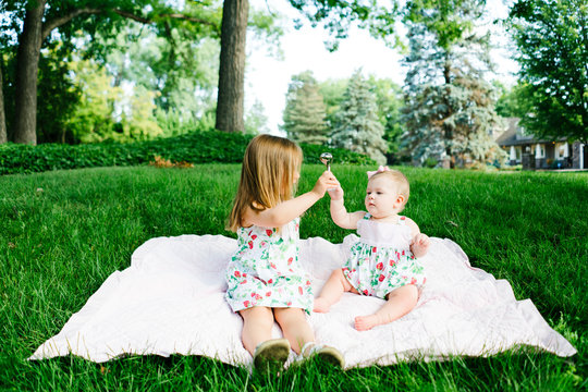 A girl gives her sister a toy while sitting on a blanket outside