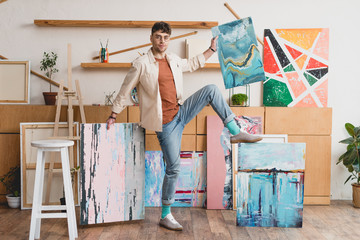 handsome artist in pink shirt and blue jeans standing in painting studio with multicolored abstract paintings