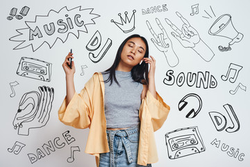 Fototapeta premium Enjoying her favourite song. Attractive and young asian woman in casual wear listening music with closed eyes while standing against grey background with music theme doodles.