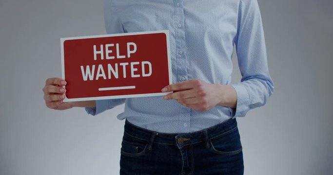 FIXED Attractive Caucasian female HR manager holding recruitment sign saying Help Wanted. Light grey background. 4K UHD