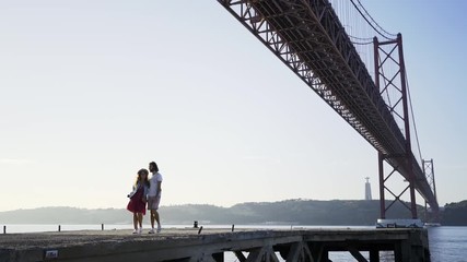 View at distance of embracing, smiling man and woman walking on concrete seafront under modern bridge in sunshine - Powered by Adobe