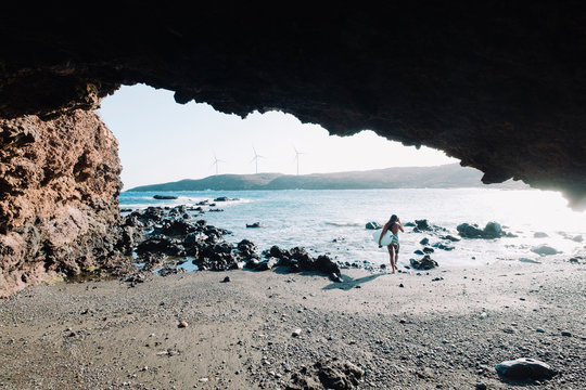View from cave of woman in bikini walking with her surfboard on beach, Tenerife, Canary Islands, Spain