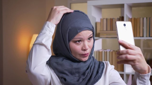 Closeup shoot of adult muslim female employee in hijab taking selfies on the phone and posing in front of the camera on the workplace indoors in the office