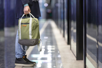 Obraz premium Closeup of man waiting train at metro station, holding a stylish light green cloth backpack, soft focus, empty subway platform on background, side view. Hand luggage concept