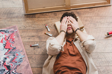 Obraz premium overhead view of exhausted artist lying on wooden floor and holding hands on face