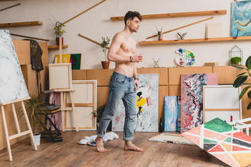 Fototapeta premium handsome half-naked artist in blue jeans standing in spacious light painting studio