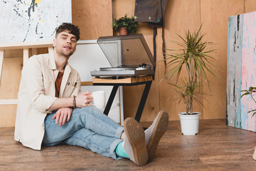handsome artist holding coffee cup and looking at camera while sitting near vinyl record  player
