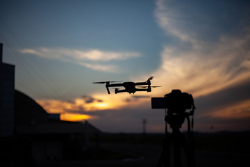 Fototapeta na wymiar Silhueta de câmera fotográfica no tripé e drone capturando o por do sol, céu azul e laranja, com nuvens.