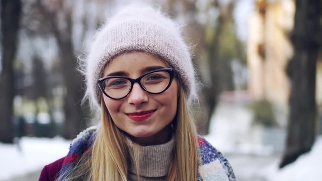Portrait of young beautiful stylish girl wearing cool knitted winter hat and glasses looking at camera smile feel happy city landscape woman outdoor holiday close up slow motion